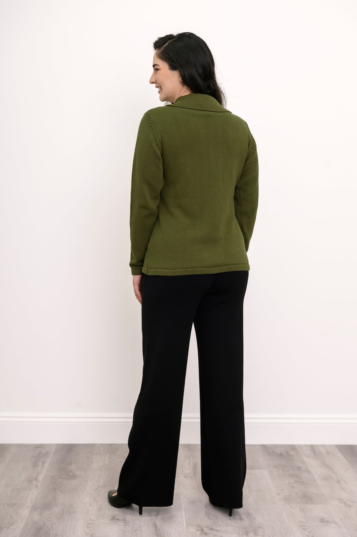 Back view of a woman wearing a hip length green cotton sweater highlighting the clean tailoring and shoulder fit.