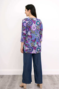 Full body back view of woman wearing long sleeve purple mosaic patterned top and blue pants