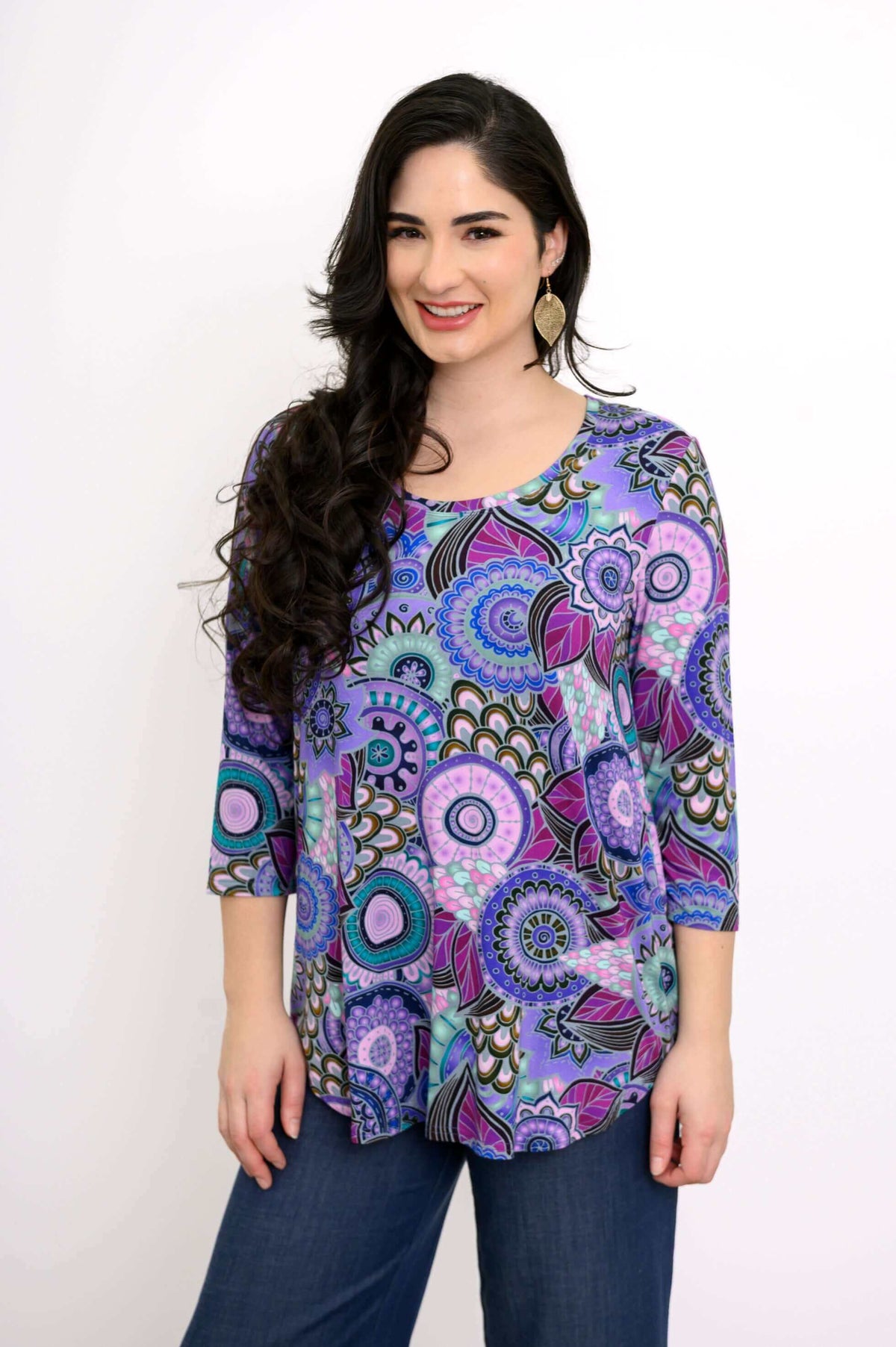 Close-up front view of woman wearing gold leaf earrings, purple top with rounded hem, and denim pants