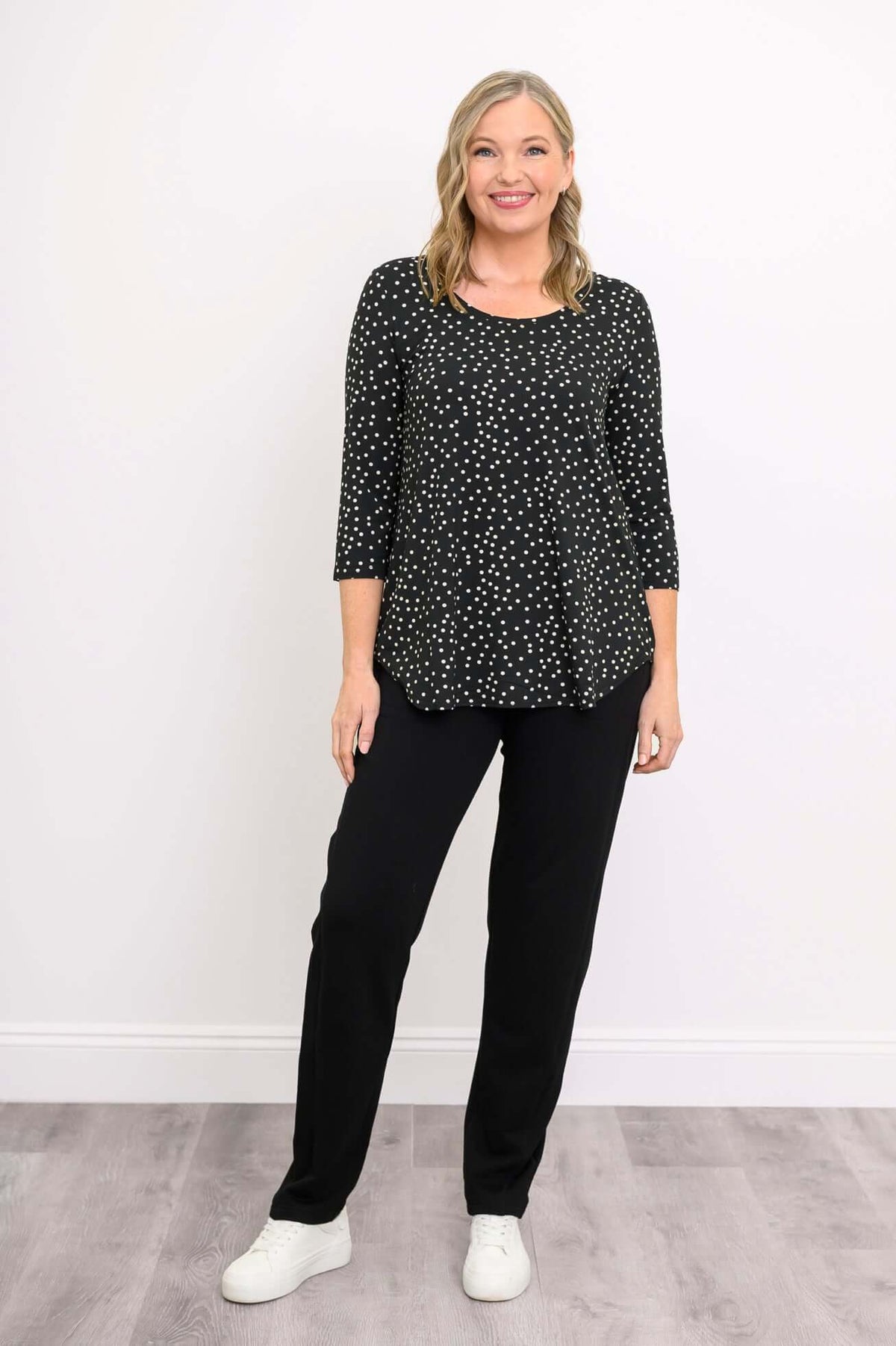 Smiling woman in black polka dot 3/4 sleeve top, black pants, and white sneakers