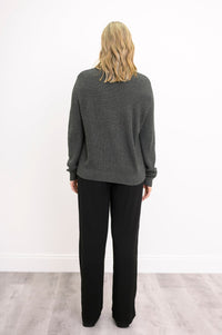Back view of a female model wearing a charcoal-colored cable-knit sweater and black pants, against a white wall and gray floor