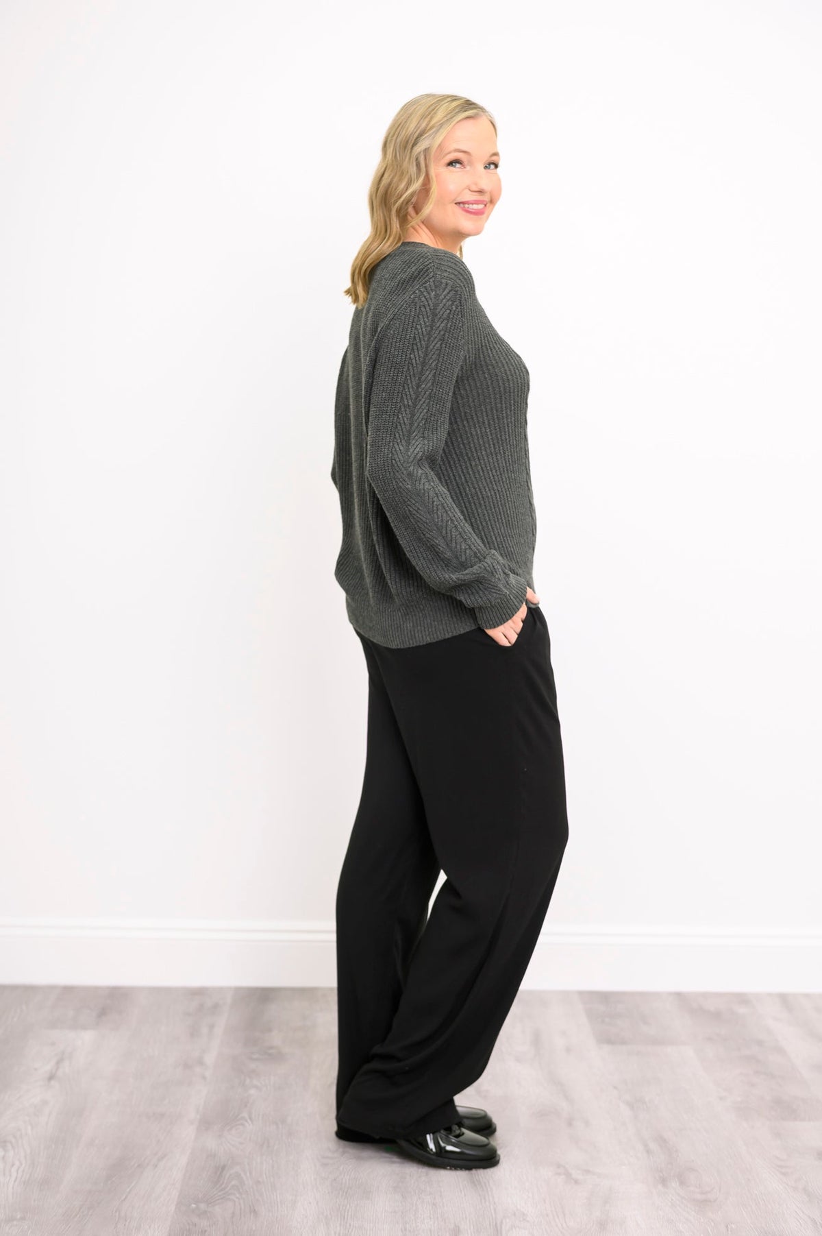Side-angle view of a female model wearing a charcoal-colored knit sweater and black pants, against a white wall and gray floor