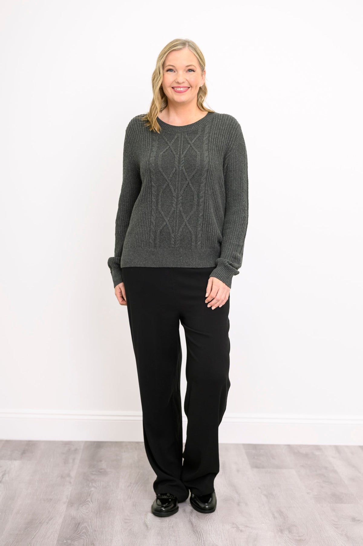Front view of a female model wearing a charcoal  long-sleeve sweater against a white wall and gray floor