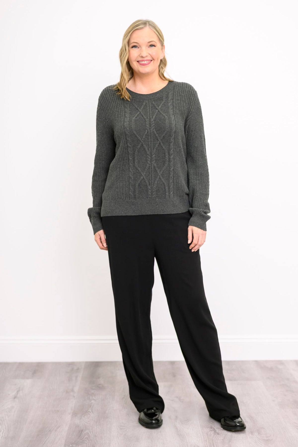 Front view of a female model wearing a graphite patterned sweater and black pants against a white wall and gray floor