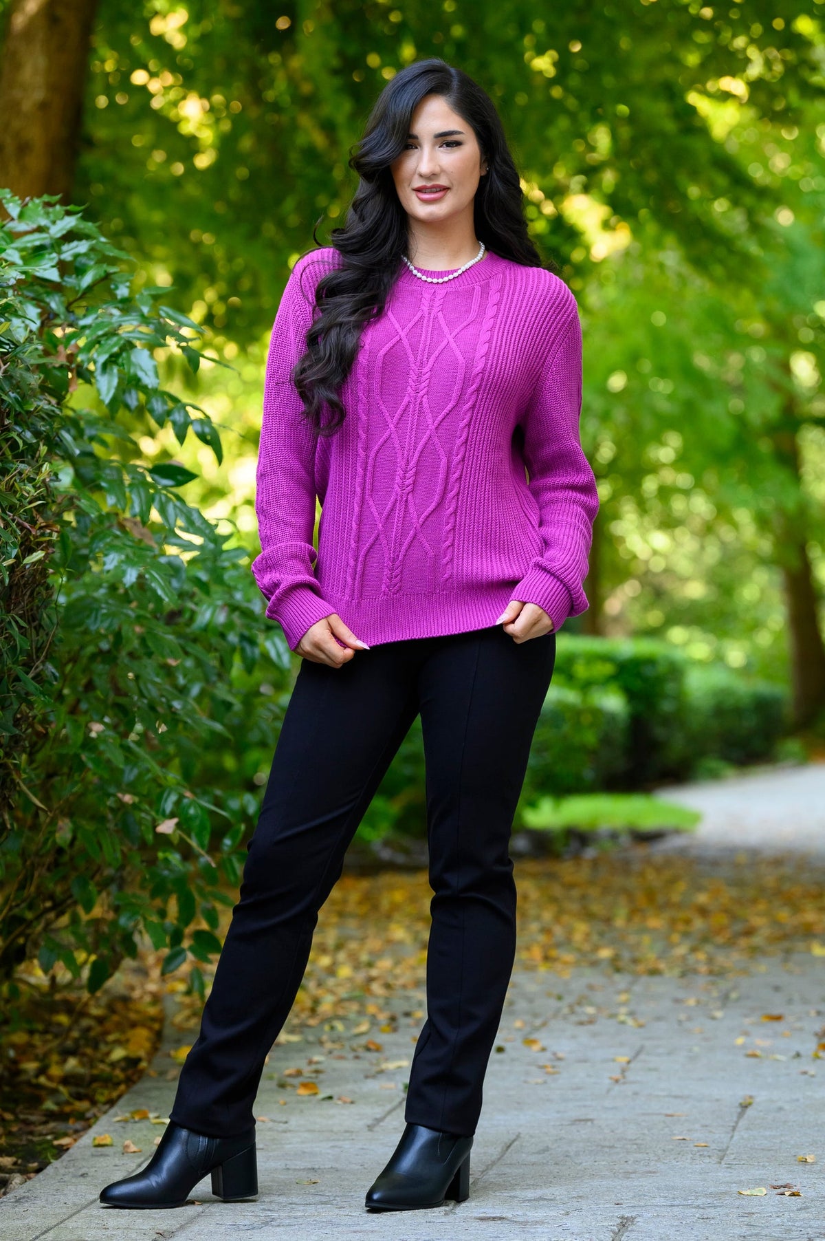 Front view of female model standing on a paved path in a park wearing a bright purple cable-knit sweater, black pants, and black ankle boots.