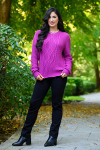 Front view of female model standing on a paved path in a park wearing a bright purple cable-knit sweater, black pants, and black ankle boots.