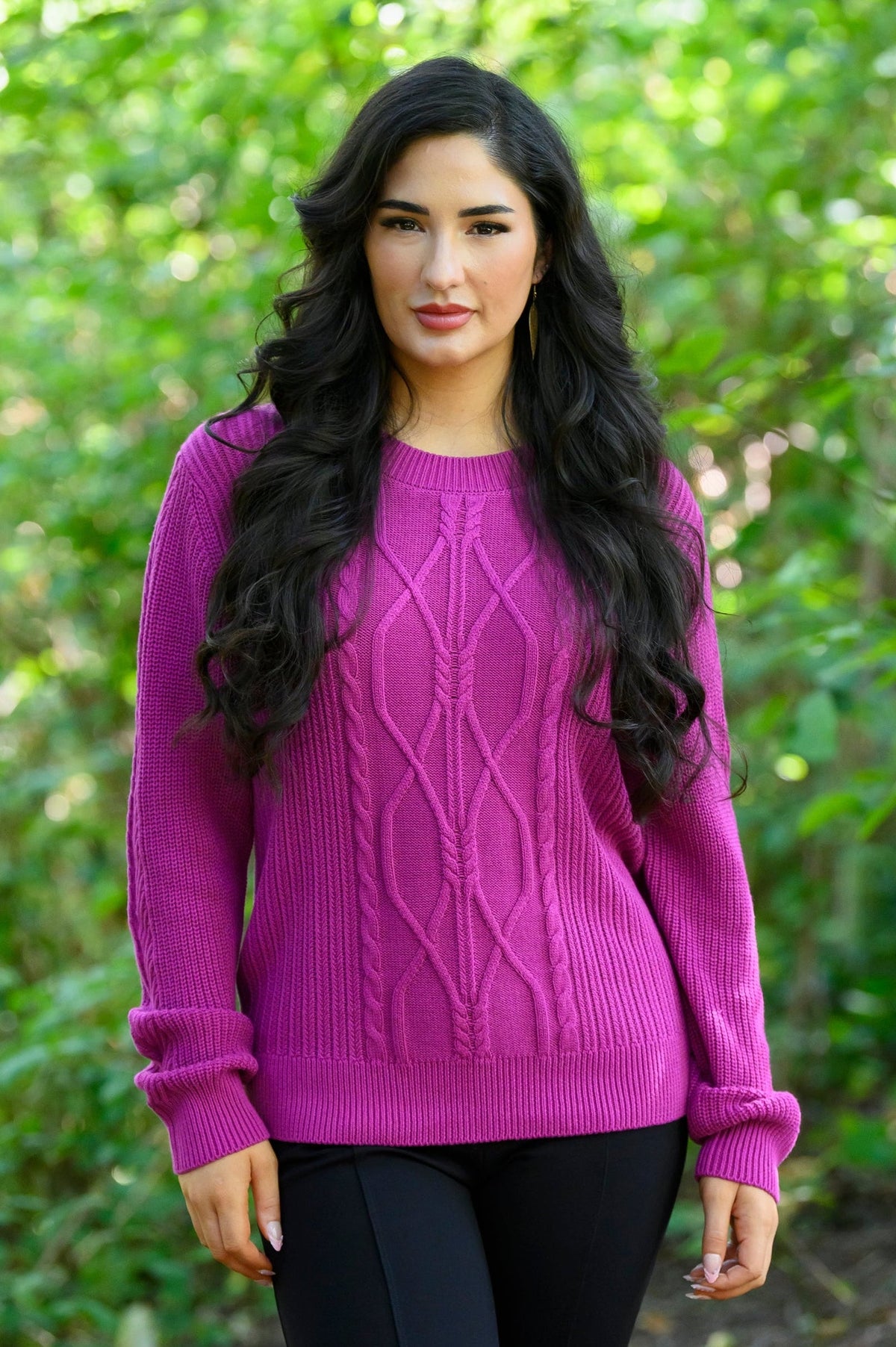 Female model wearing a bright magenta cable-knit sweater and black pants, standing outdoors in front of a blurred green forest background.