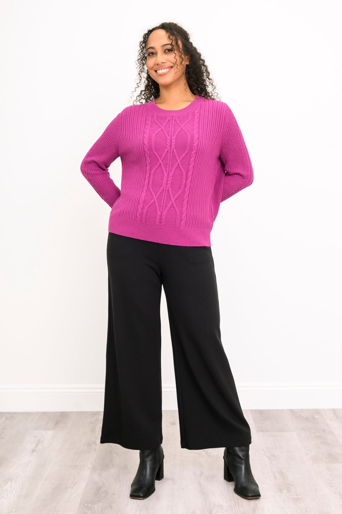 Front view of a female model wearing a vibrant rose-pink sweater and black trousers, against a white wall and gray floor