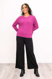 Front view of a female model wearing a vibrant rose-pink sweater and black trousers, against a white wall and gray floor