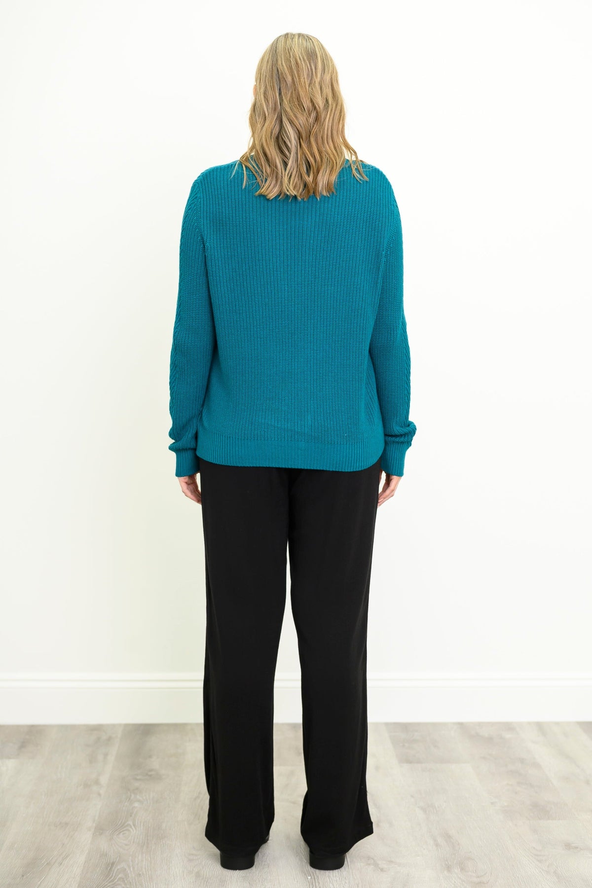 Back view of female model wearing a teal ribbed knit sweater and black wide-leg pants, standing against a white wall on a light gray wood floor.