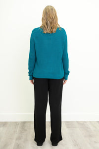 Back view of female model wearing a teal ribbed knit sweater and black wide-leg pants, standing against a white wall on a light gray wood floor.