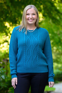 Front view of female model standing on a stone path in a green park, wearing a teal cable-knit sweater, black pants, and black ankle boots.