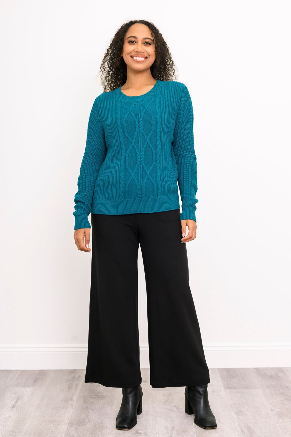 Front view of a female model wearing a dark turquoise knit top and loose pants, against a white wall and gray floor