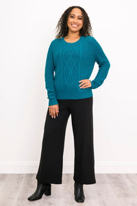 Front view of female model wearing a teal long-sleeve cable-knit sweater paired with black wide-leg pants and black ankle boots, standing against a white wall and gray floor