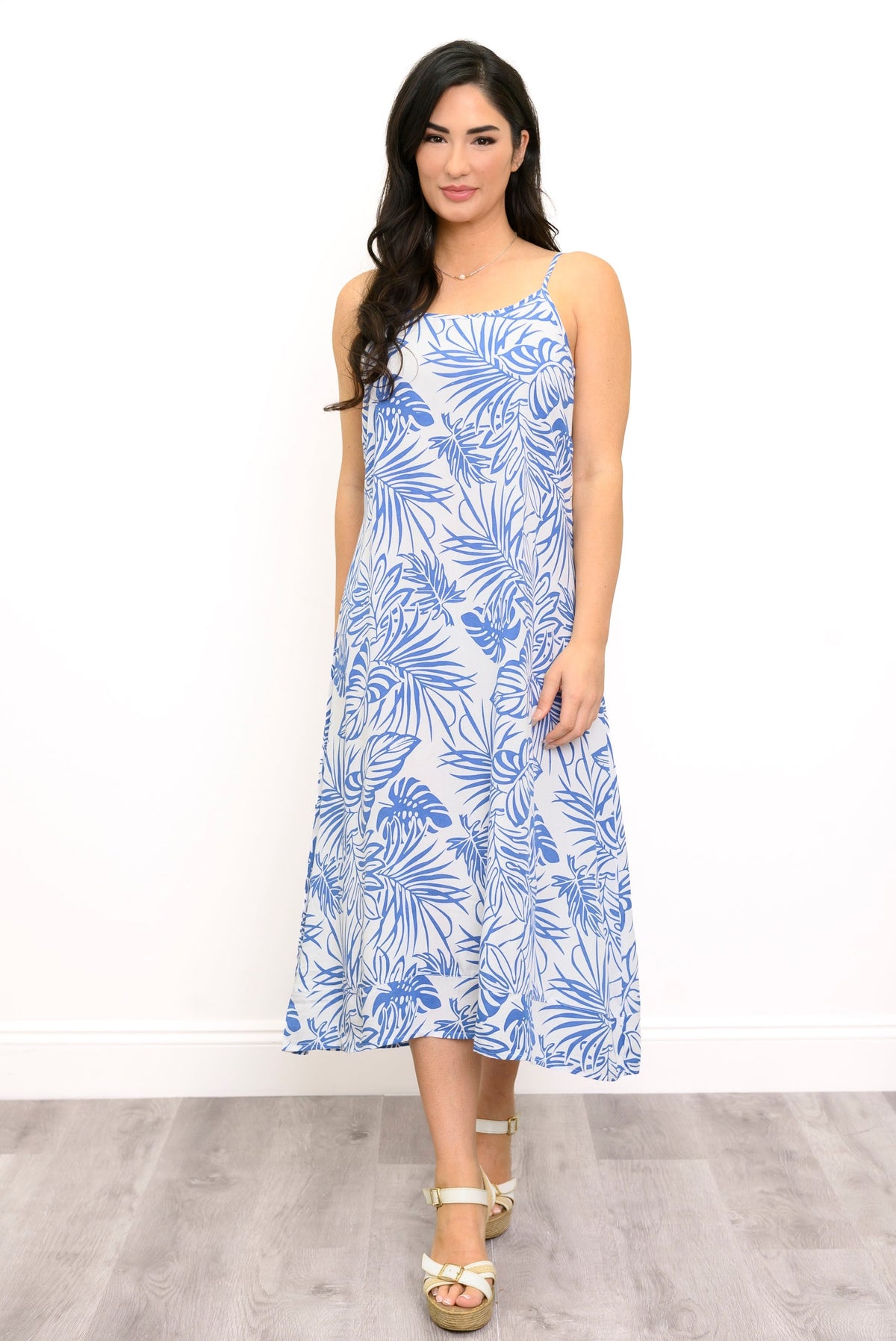 A woman modeling a sleeveless midi dress with a blue tropical palm leaf pattern on a white background, paired with white platform sandals, set against a plain white wall and light wood floor.