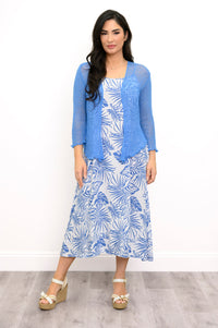 A woman modeling a three-piece blue and white tropical print outfit, consisting of a blue lace cardigan, a white midi dress with blue palm leaf print, and beige wedge sandals, set against a plain white wall and light wood floor.