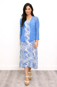 A woman modeling a light blue lace cardigan over a white midi dress with a blue tropical palm leaf print, standing against a white wall on a gray wood floor.