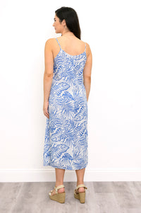 A woman wearing a sleeveless midi slip dress with a blue tropical leaf pattern on a white background, shown from the back against a white wall.