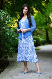 A woman stands on a paved garden path wearing a blue and white tropical print midi dress with a sheer blue lace cardigan and blue pointed shoes, set against a lush green background.