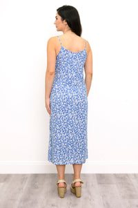 A woman stands with her back to the camera wearing a sleeveless midi dress with a blue and white floral pattern, paired with beige wedge sandals, set against a plain white wall and light gray wood floor.