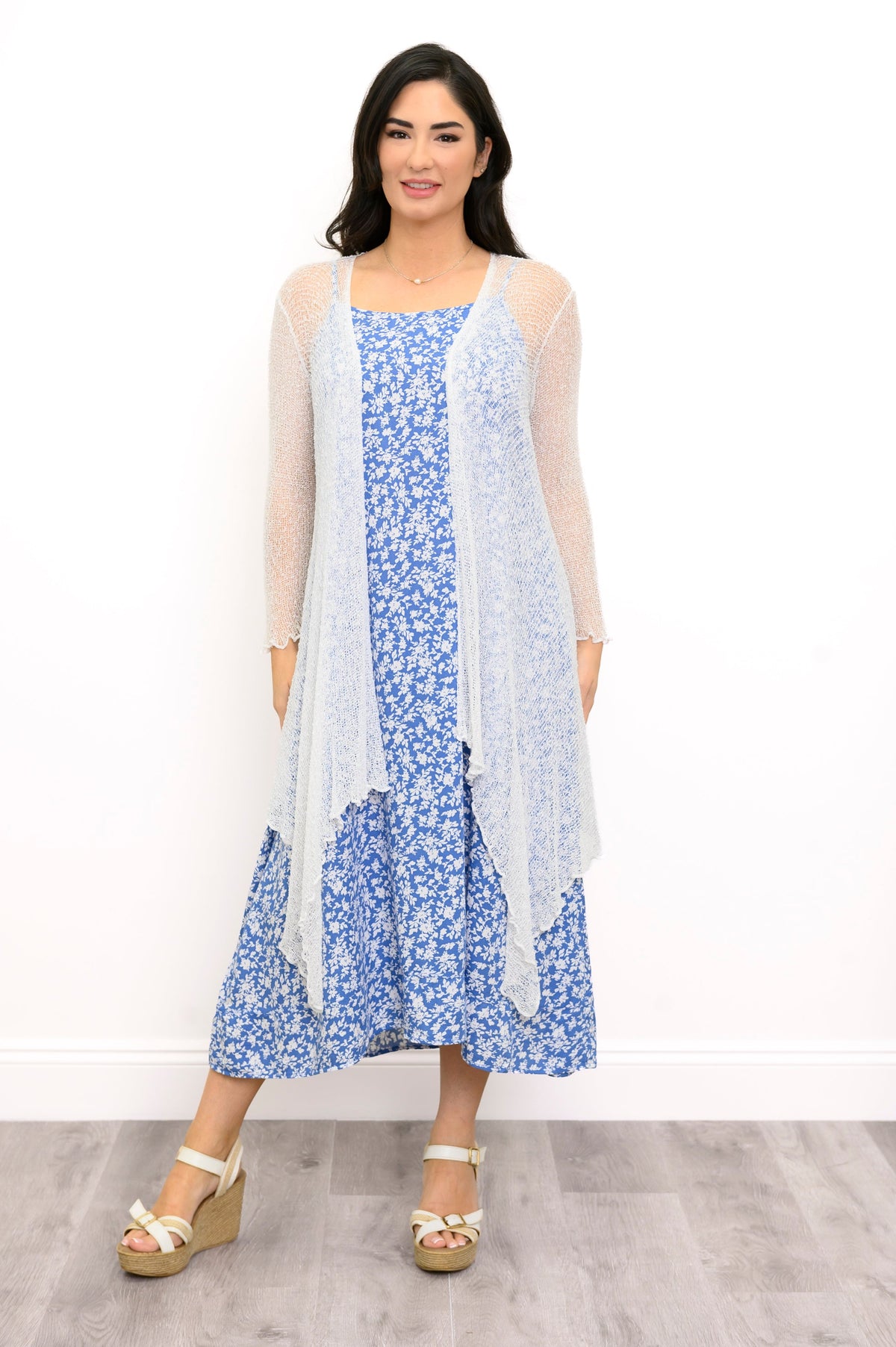 A woman modeling a light blue and white floral midi dress with a sheer white long-sleeve cardigan and beige wedge sandals, standing against a white wall.