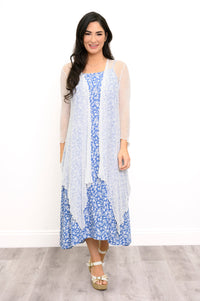 A woman modeling a blue and white floral midi dress with a sheer white long-sleeve cardigan and white wedge sandals, standing against a white wall.