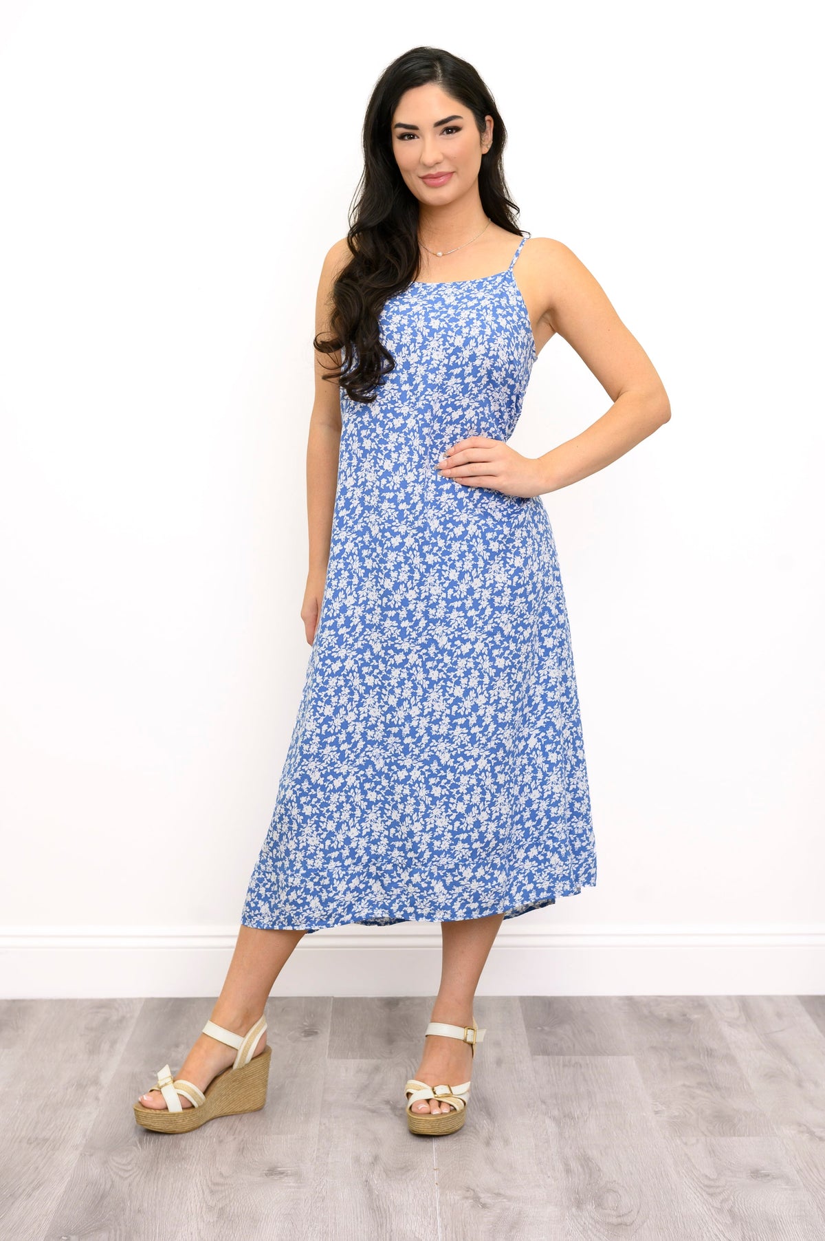 A woman modeling a sleeveless midi dress with a blue and white floral pattern, paired with beige wedge sandals, standing against a white wall and light gray wood floor.