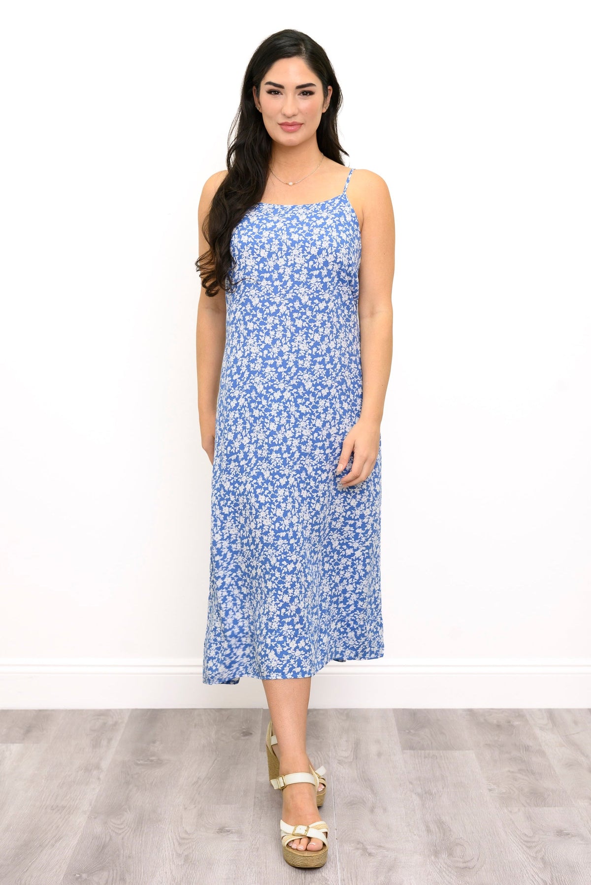 A woman modeling a sleeveless midi dress with a blue and white floral pattern, standing against a white wall on a light gray wood floor.