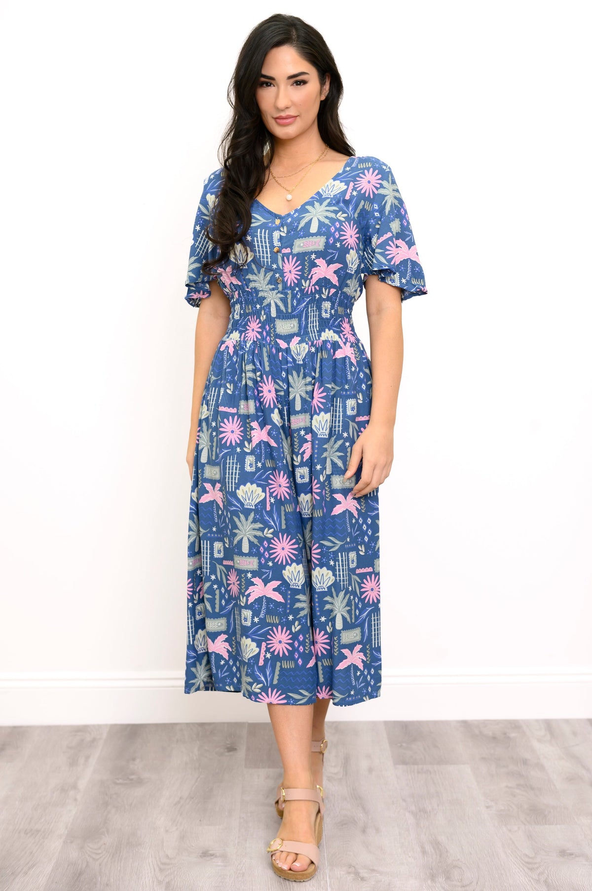 A woman modeling a short-sleeve midi dress with a blue, pink, and white tropical floral print, standing against a white wall and light gray wood floor.