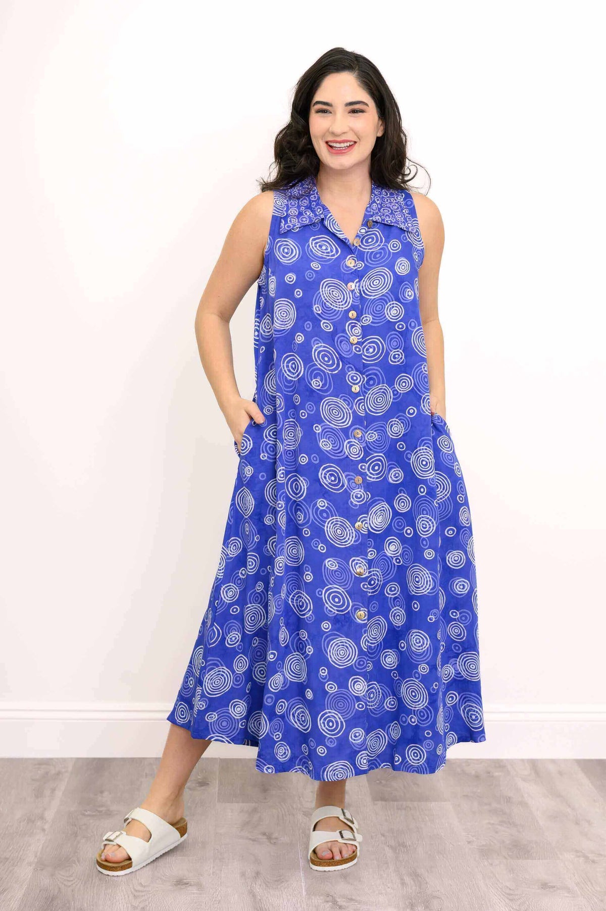 Model smiling with hands in pockets, showcasing a blue sleeveless dress with bold swirl patterns