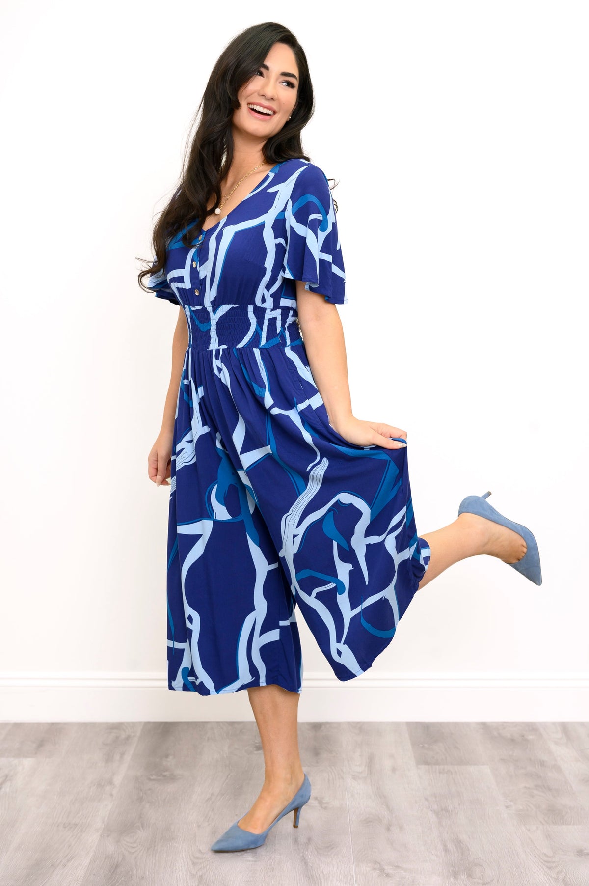A woman modeling a blue and white abstract print midi jumpsuit, paired with blue pointed-toe heels, standing against a white wall and light wood floor.