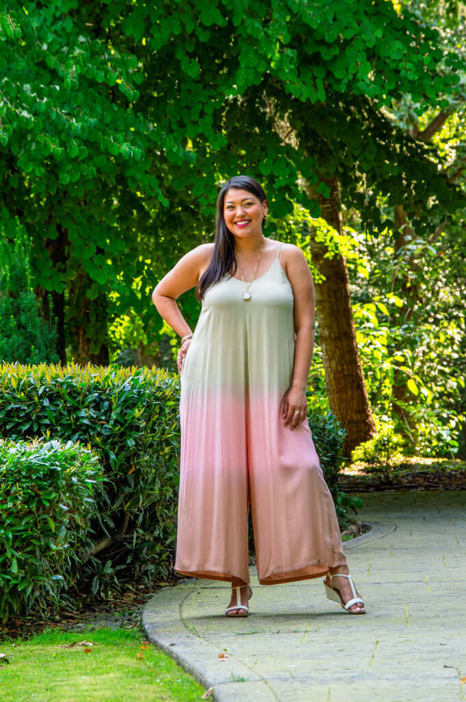 Woman in blush ombre jumpsuit standing on a garden path surrounded by lush greenery