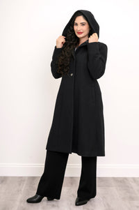 Woman posing with her coat hood on in a black loose-fitting coat and black pants