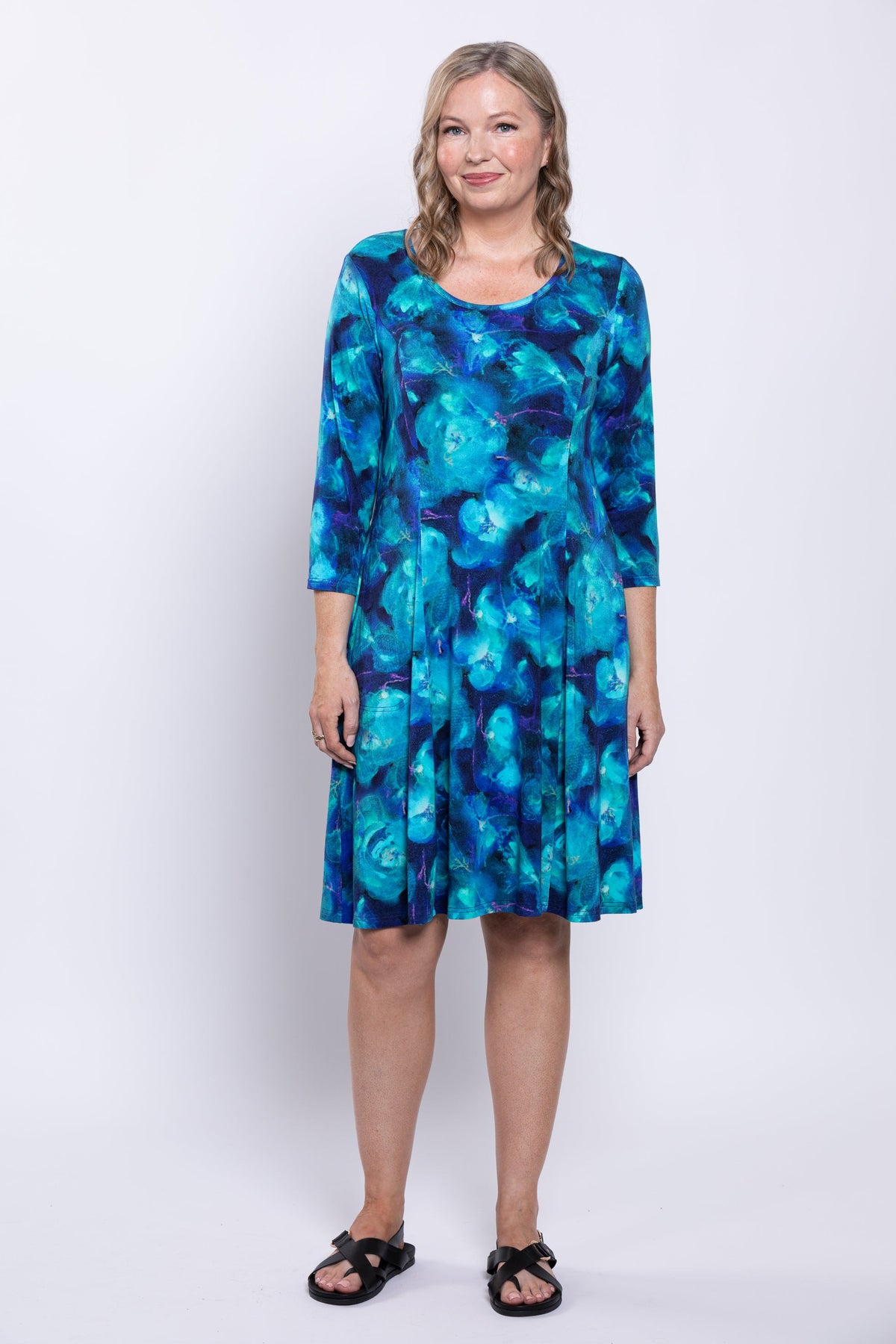 Woman modeling with her hands to her side and smiling as she wears a blue dress and black sandals