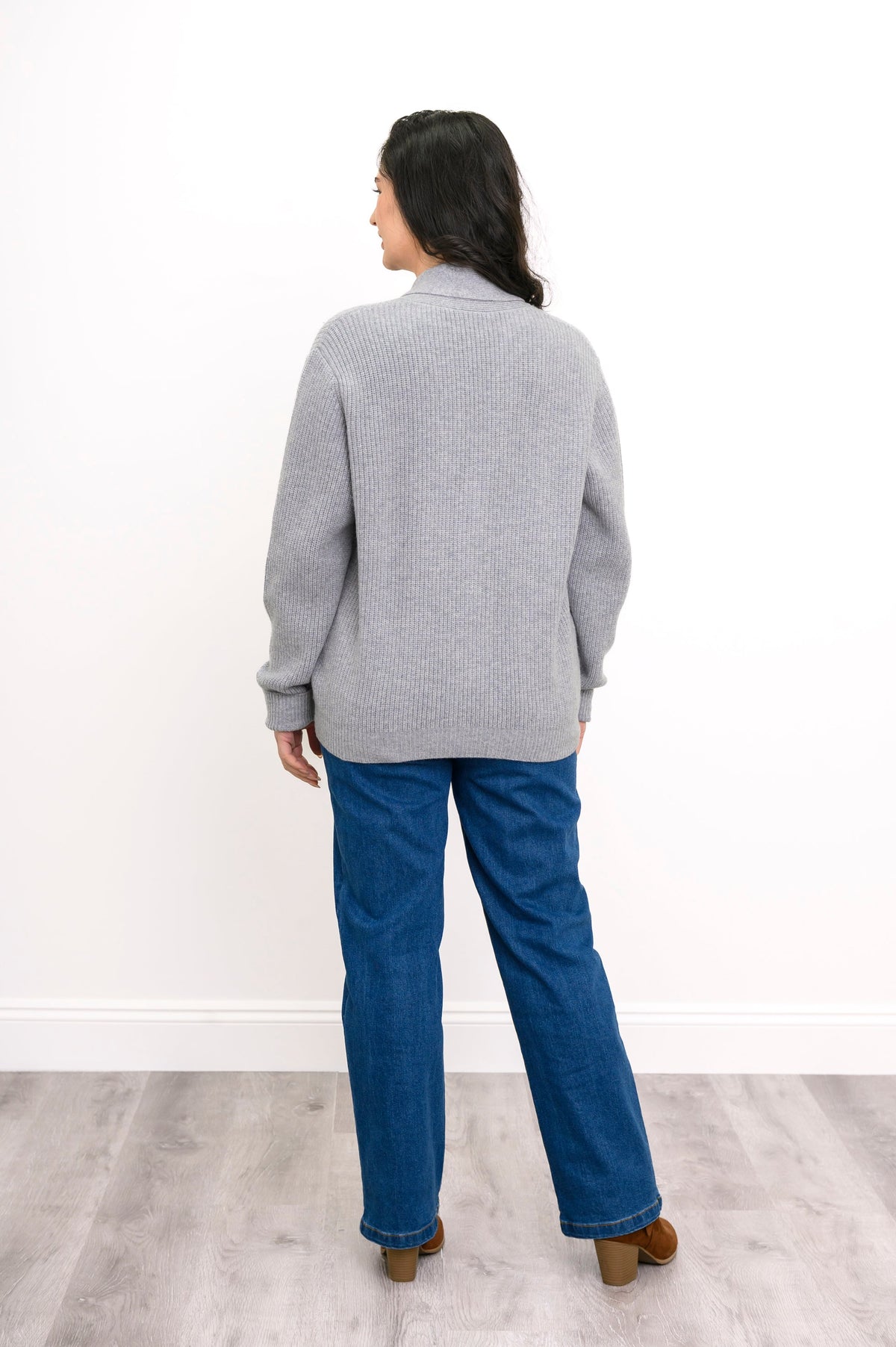 Back view of woman wearing pale grey chunky knit sweater with denim pants and brown heeled boots
