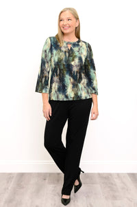 Front view of a female model wearing a watercolor green top with long sleeves with black pants and heels, looking to the side, against a white wall and gray floor