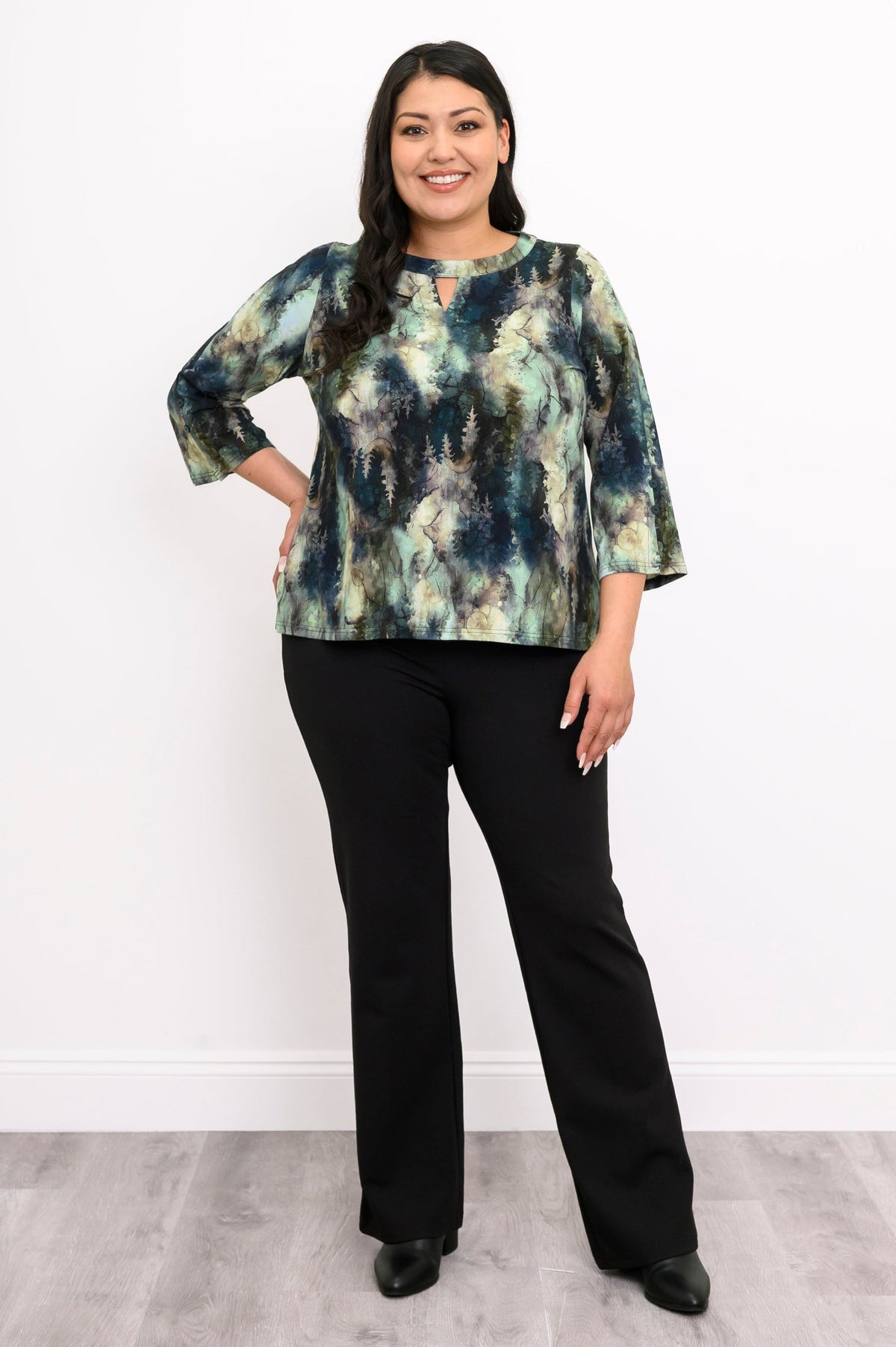 Front view of a female model wearing a watercolor green top with long sleeves with black pants and heels, against a white wall and gray floor