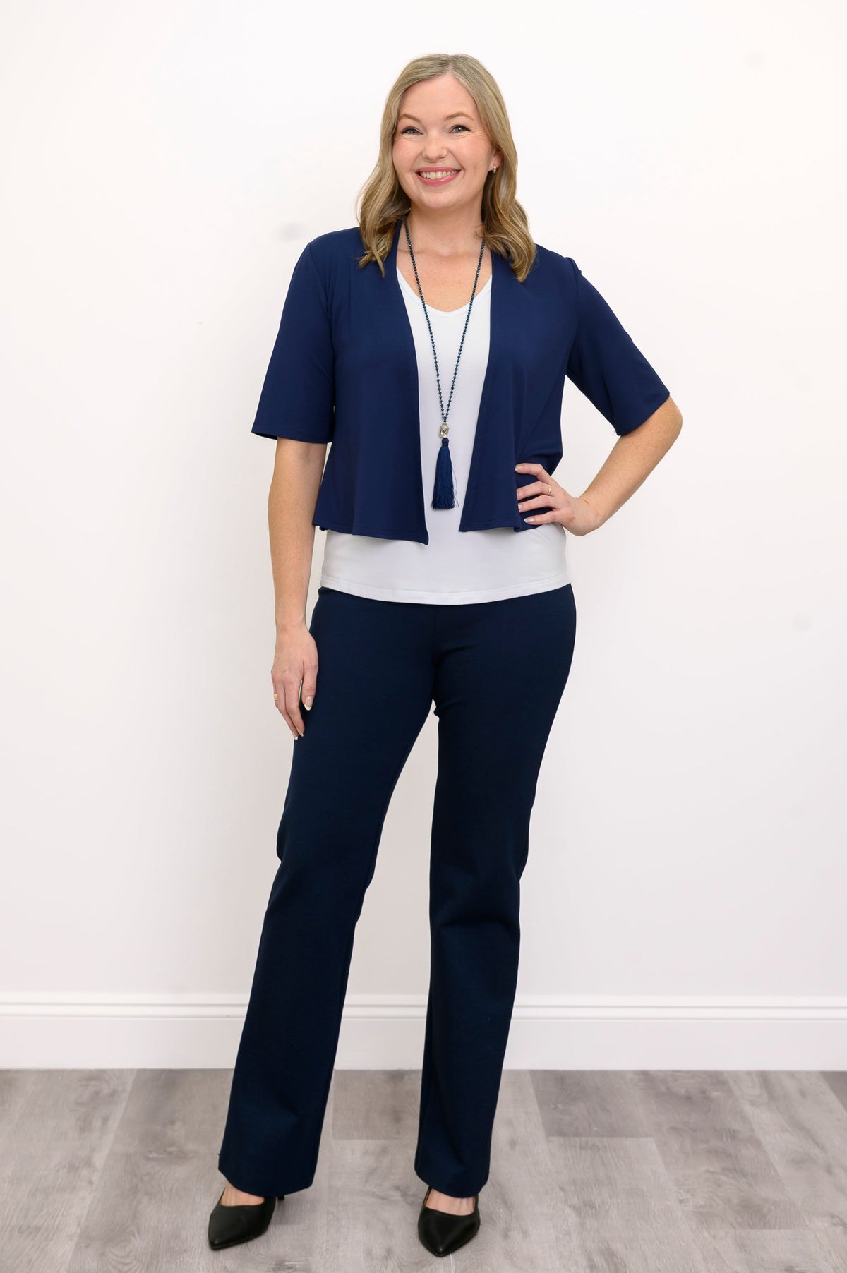 Front view, woman wearing navy blue jacket, white top, blue necklace, and blue pants against a white background