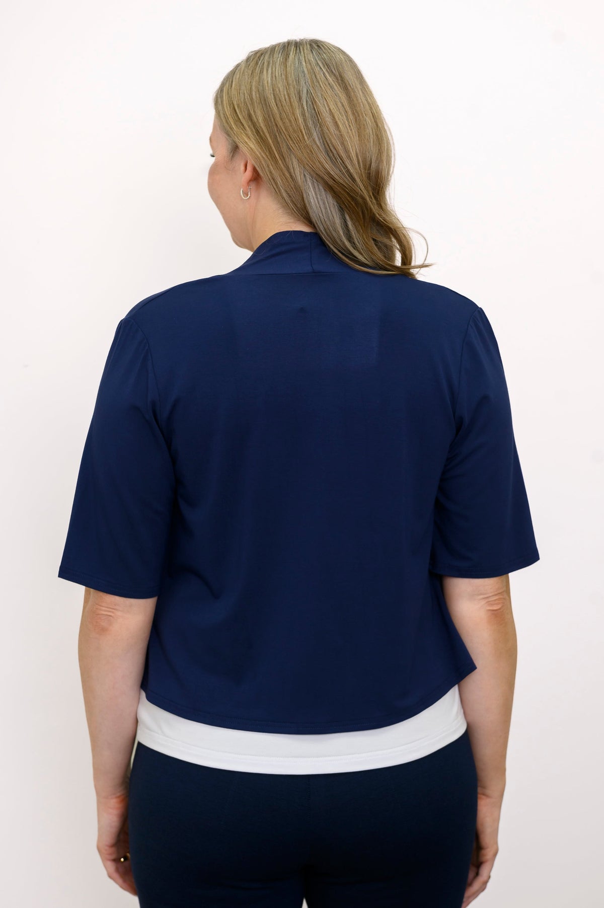Back view, woman wearing navy blue jacket, white top and blue pants against a white background