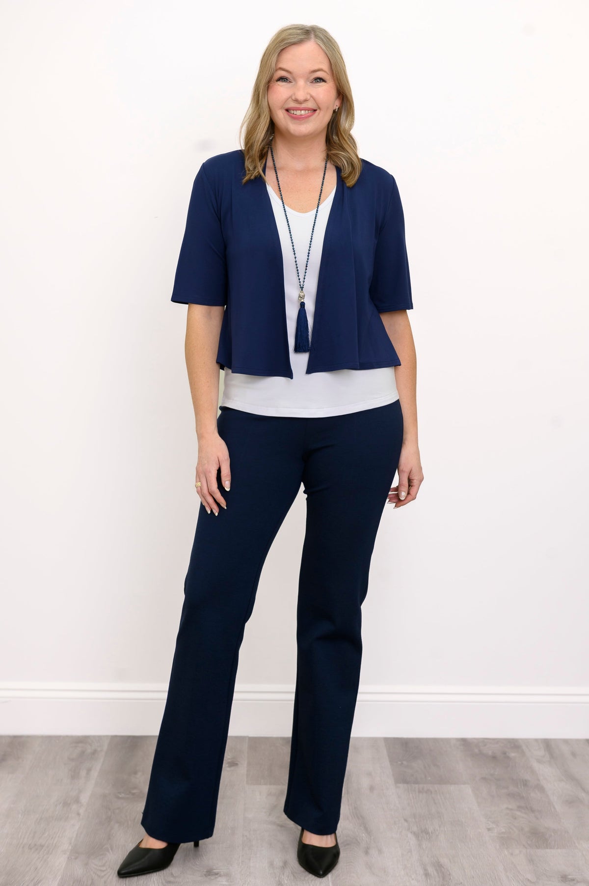 Front view, woman wearing navy blue jacket, white top, blue necklace, and blue pants against a white background