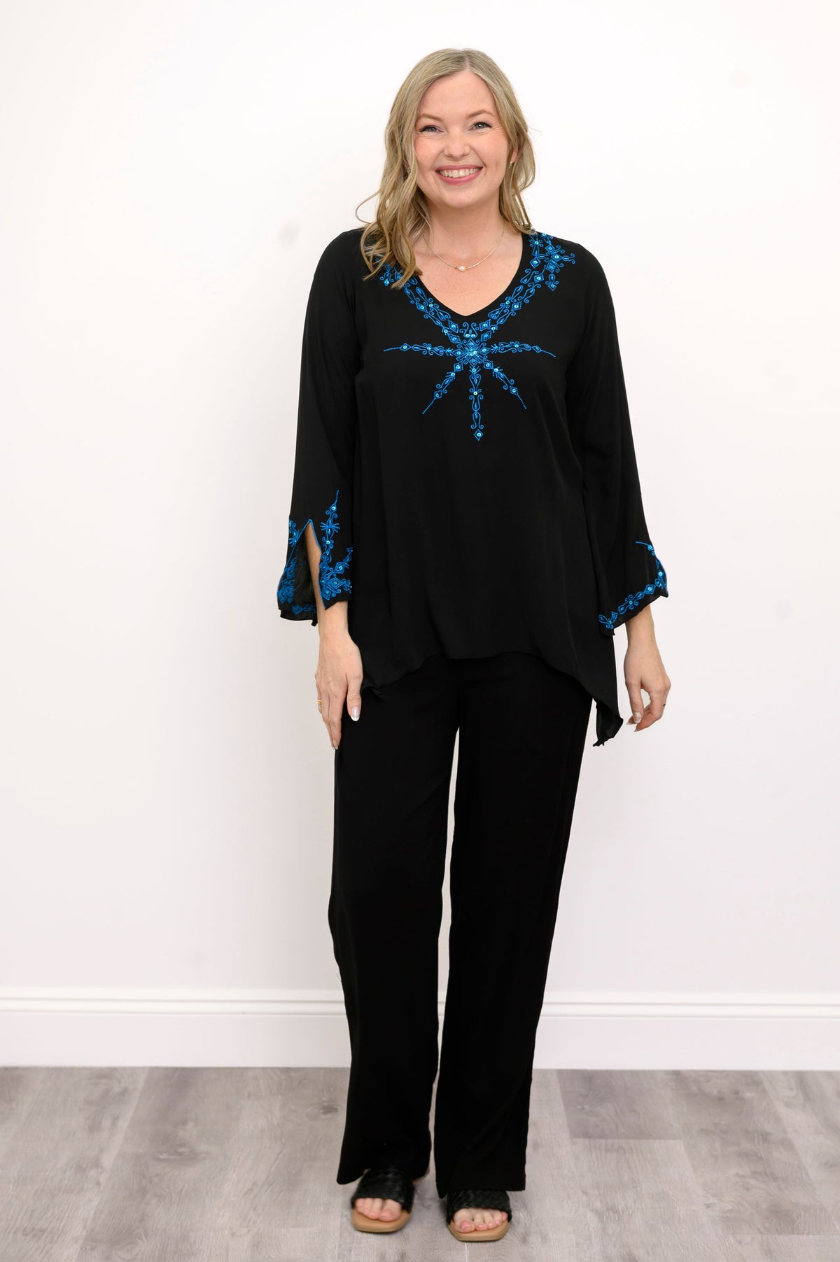 Front view of woman wearing a black top with blue embellishments and black pants against a white background