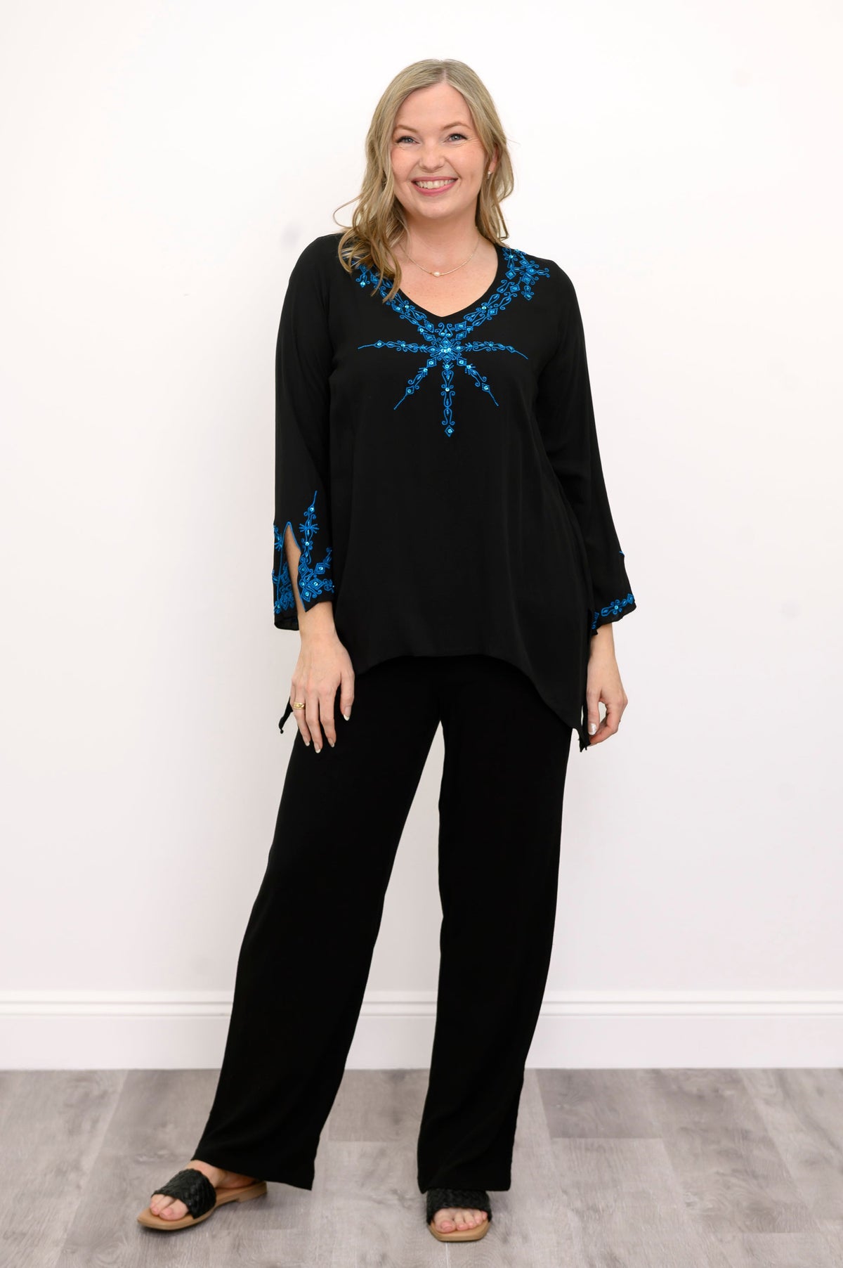Front view of woman wearing a black top with blue embellishments and black pants against a white background