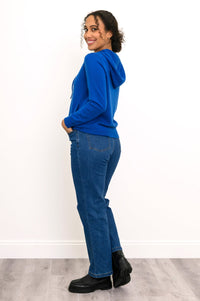 Woman wearing a knit bright blue hoodie with pockets and denim pants in front of a white background, side view