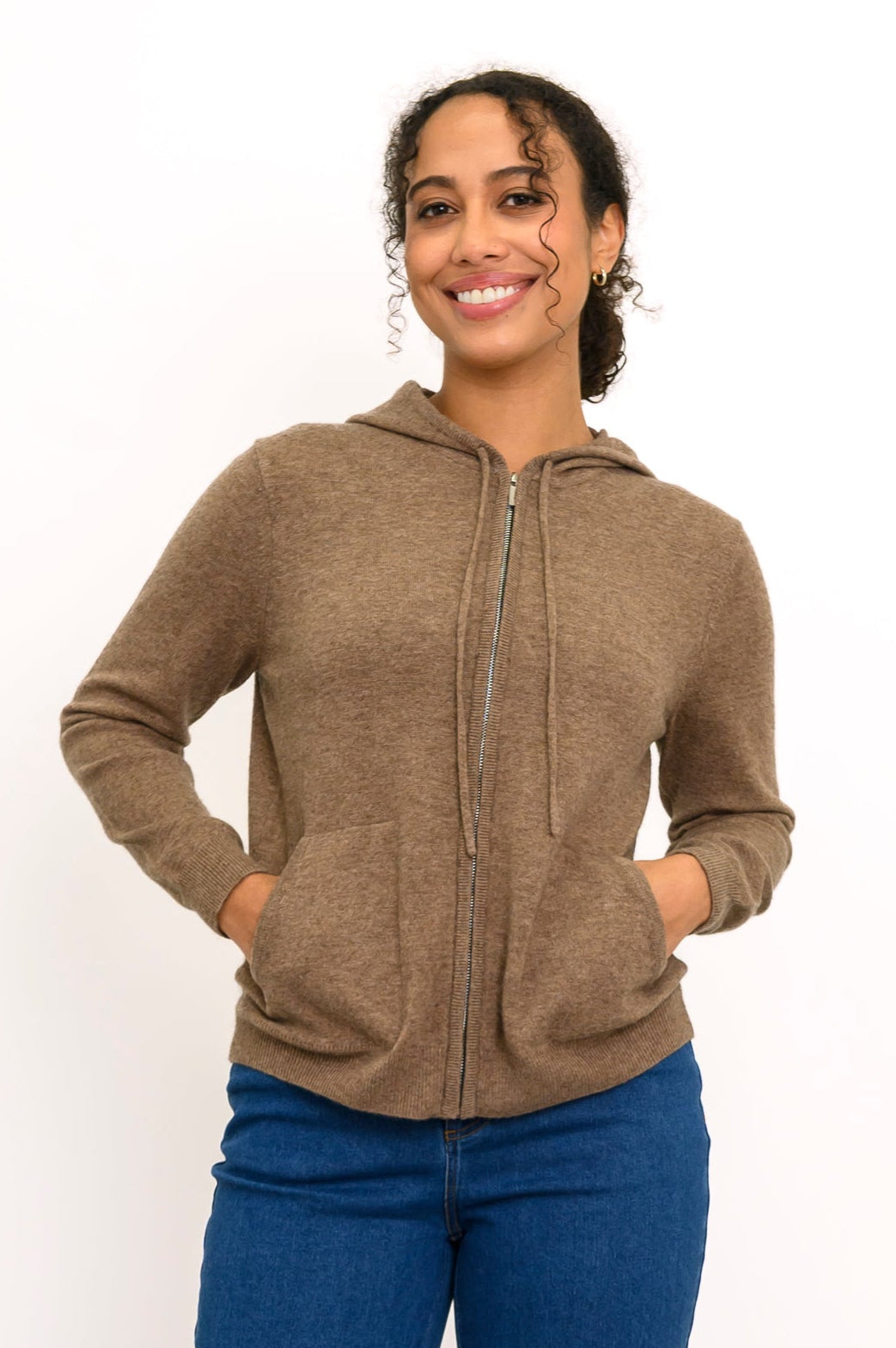 Close-up front view of woman smiling with her hands in the pockets of her light brown knit hoodie