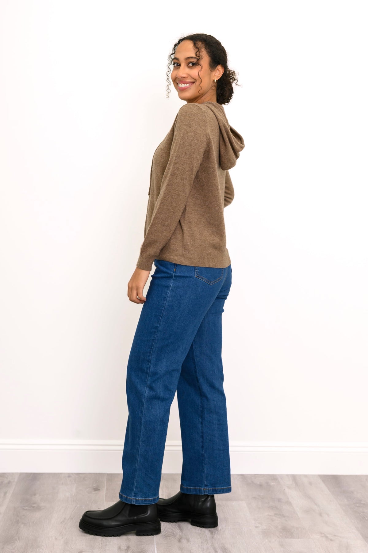 Woman wearing a knit brown hoodie with pockets and denim pants in front of a white background, side view