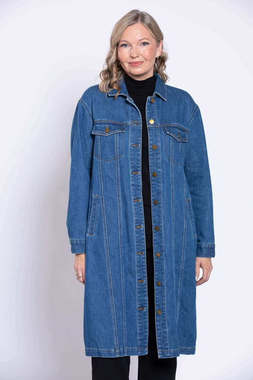 Front view of a woman wearing a longline blue denim trucker jacket over an all black outfit on white.