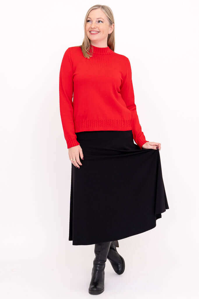 Full-body view of a woman in a red sweater and black skirt, holding the skirt