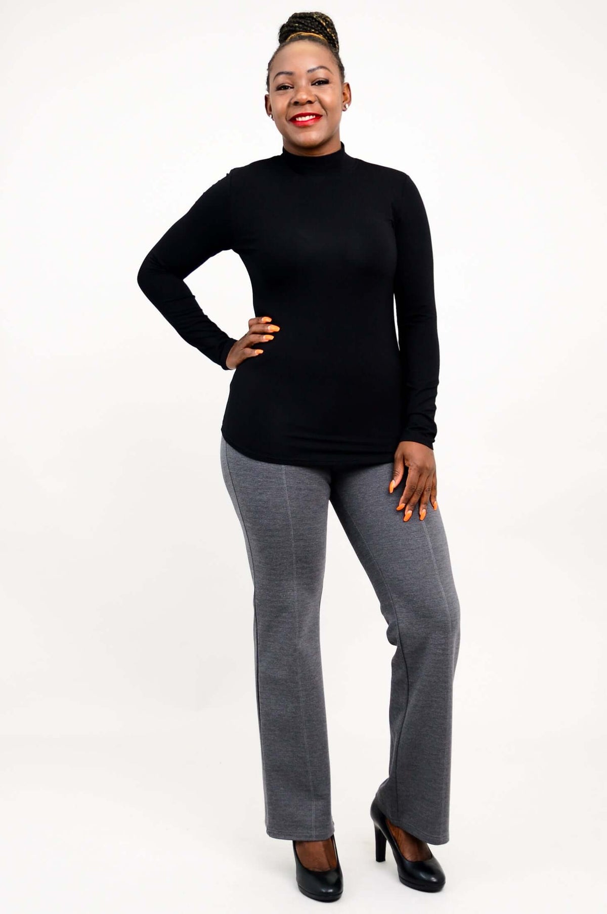 Full body view of a woman in a black fitted mock neck top and gray pants with heels