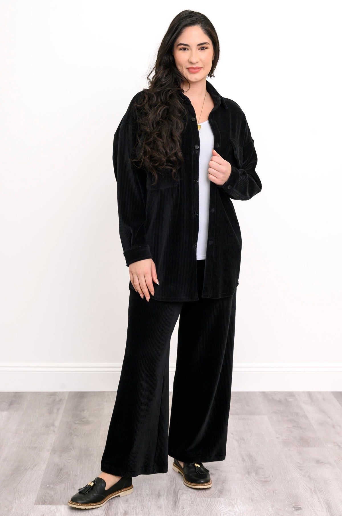 A woman modeling a black velvet two-piece lounge set consisting of a button-up shirt jacket and matching wide-leg pants, styled over a white top and black loafers.