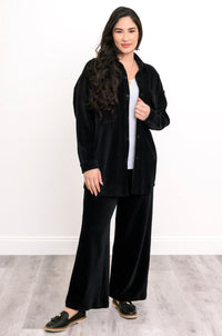 A woman modeling a black velvet two-piece lounge set consisting of a button-up shirt jacket and matching wide-leg pants, styled over a white top and black loafers.